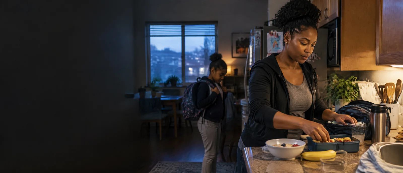 A mother prepares breakfast and a school bag before dawn while her child gets ready nearby.